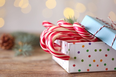 Photo of Christmas candy canes in gift box on wooden table against blurred lights, closeup. Space for text