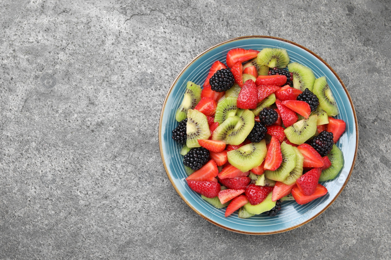 Photo of Plate of delicious fresh fruit salad on grey textured table, top view. Space for text