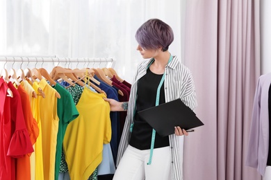 Beautiful young stylist with clipboard choosing clothes from rack in studio