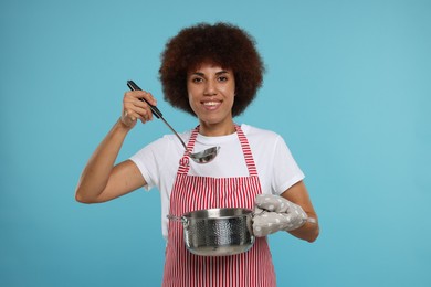 Photo of Happy young woman in apron holding ladle and cooking pot on light blue background