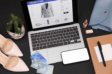 Photo of Online store website on laptop screen. Computer, smartphone, credit cards, stationery, women's shoes, bag and lipstick on black background, above view