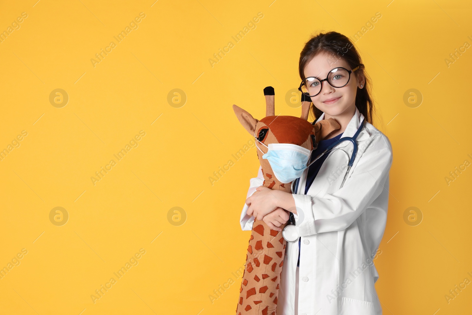 Photo of Little girl with eyeglasses and stethoscope as doctor hugging toy giraffe on yellow background, space for text. Pediatrician practice