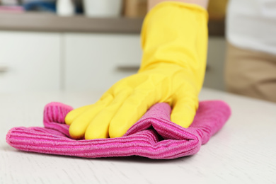 Photo of Woman in gloves wiping white wooden table with rag indoors, closeup