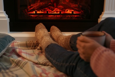 Woman in warm socks with cup of hot drink resting near fireplace at home, closeup