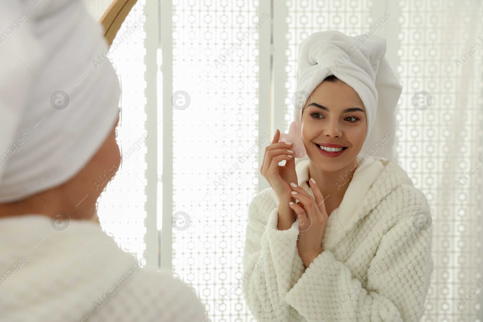 Photo of Beautiful young woman doing facial massage with gua sha tool in front of mirror at home