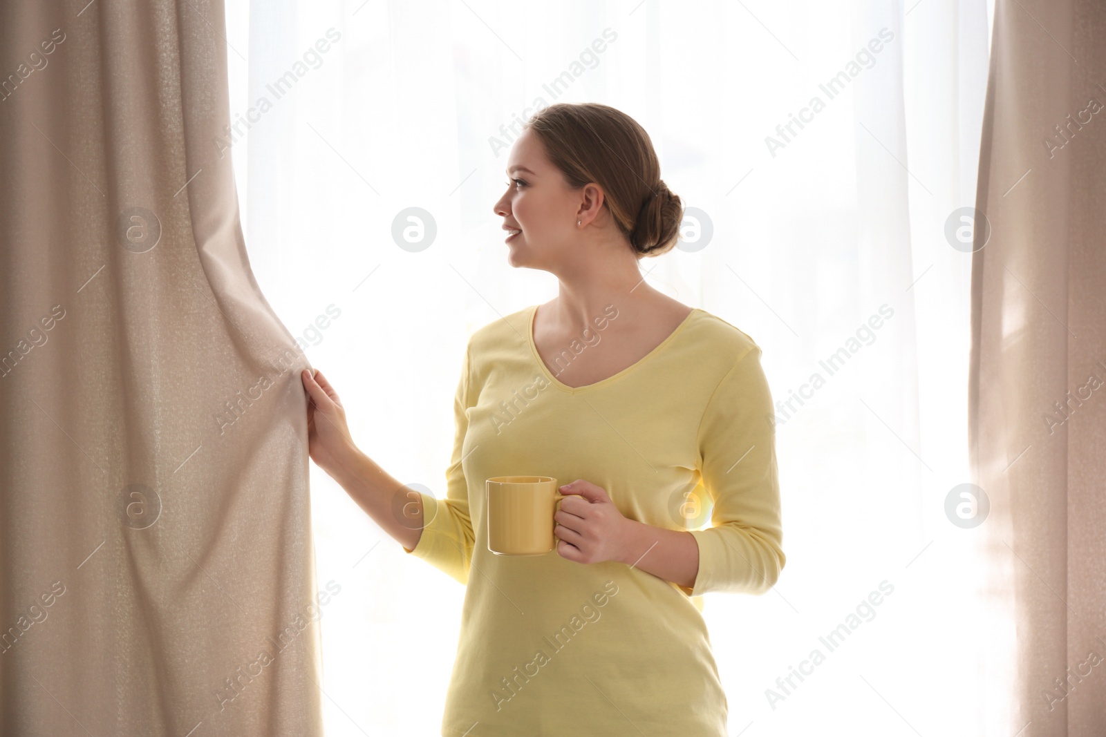 Photo of Woman with cup of coffee opening window curtain at home in morning