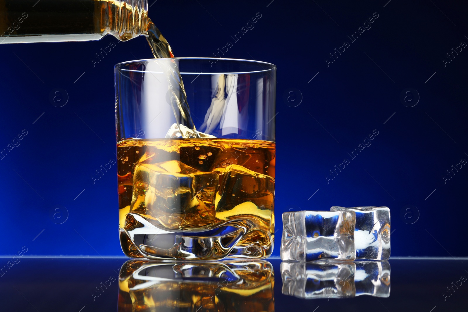 Photo of Pouring whiskey into glass with ice cubes at table against dark blue background