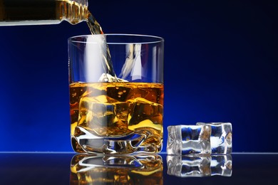 Photo of Pouring whiskey into glass with ice cubes at table against dark blue background