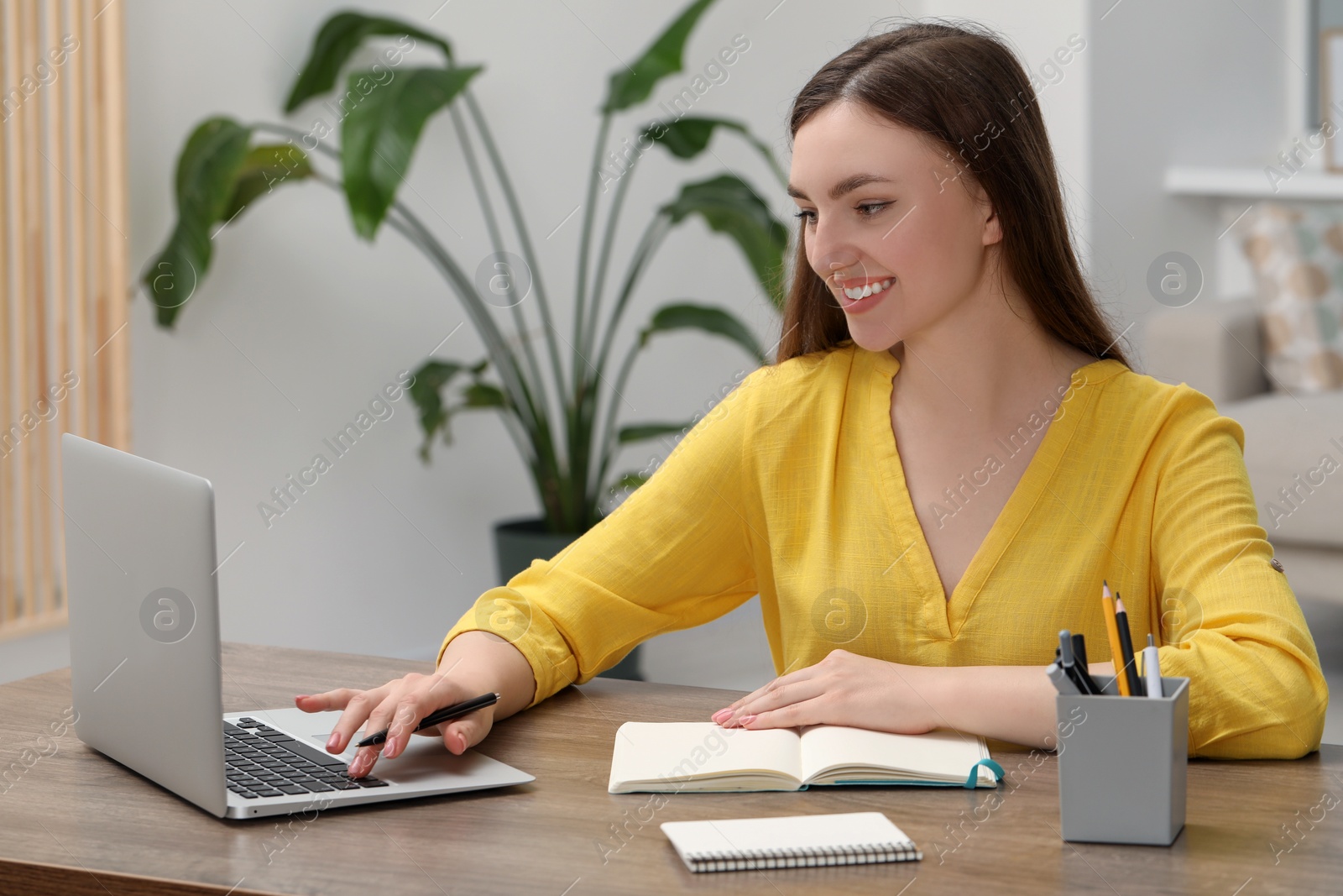 Photo of Happy young woman with notebook working on laptop at wooden table indoors
