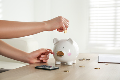 Photo of Woman putting money into piggy bank at wooden table indoors, closeup