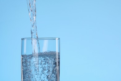 Pouring water into glass on light blue background, closeup. Space for text