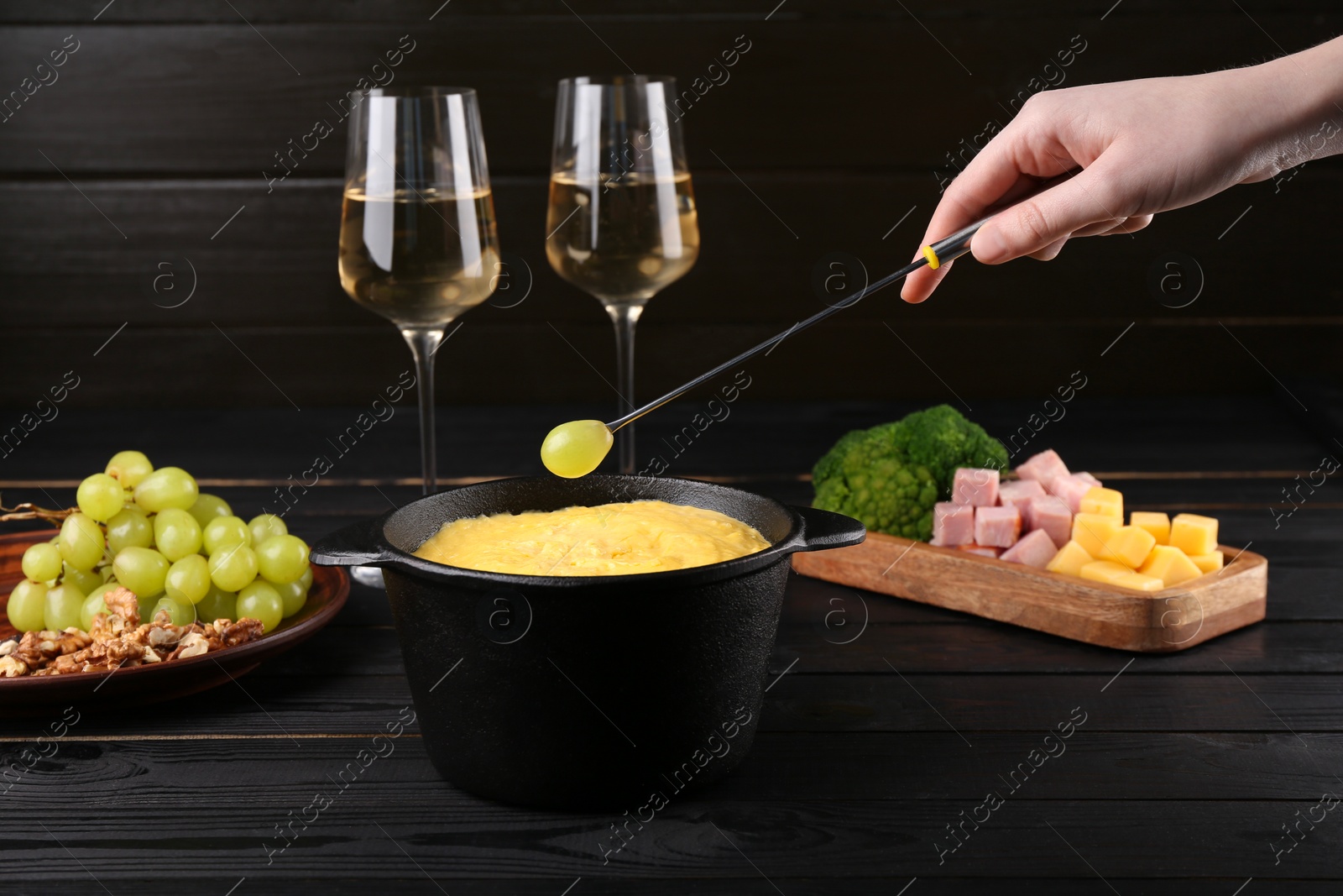 Photo of Woman dipping grape into fondue pot with melted cheese at black wooden table, closeup