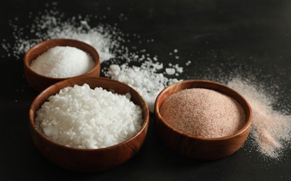 Photo of Different types of organic salt in bowls on black table, closeup
