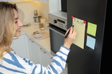 Photo of Young woman writing To do list on refrigerator door in kitchen