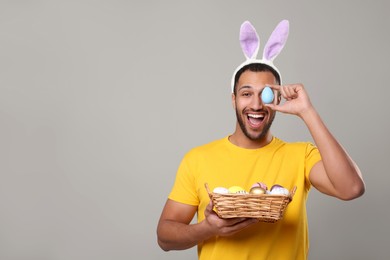 Happy African American man in bunny ears headband covering eye with Easter egg on gray background. Space for text