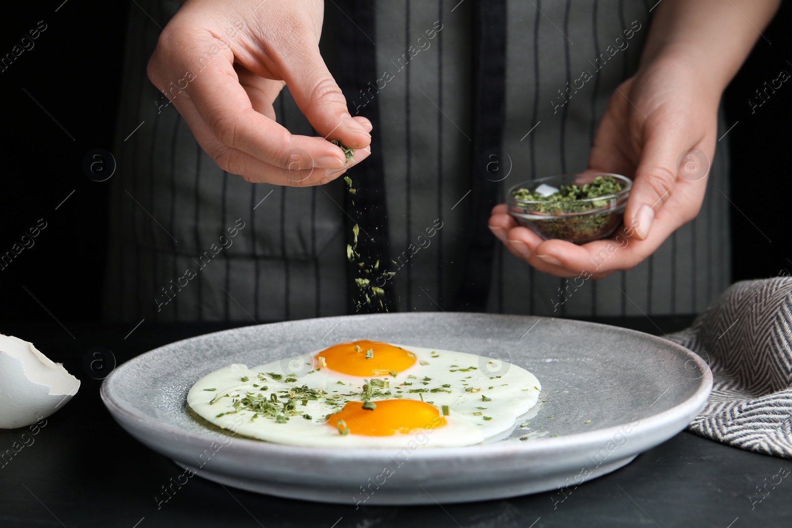 Photo of Woman adding herbs to fried eggs at black table, closeup