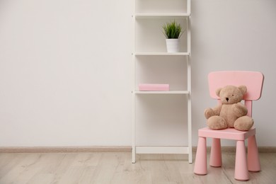 Photo of Teddy bear on chair and shelving unit near wall in child room. Space for text