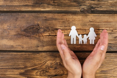 Photo of Young man protecting paper family figure with his hands on wooden background, top view. Space for text