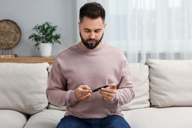 Photo of Diabetes test. Man checking blood sugar level with lancet pen on sofa at home