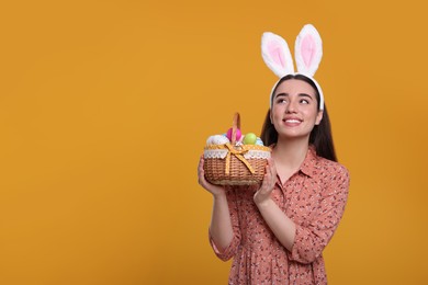 Photo of Happy woman in bunny ears headband holding wicker basket of painted Easter eggs on orange background. Space for text