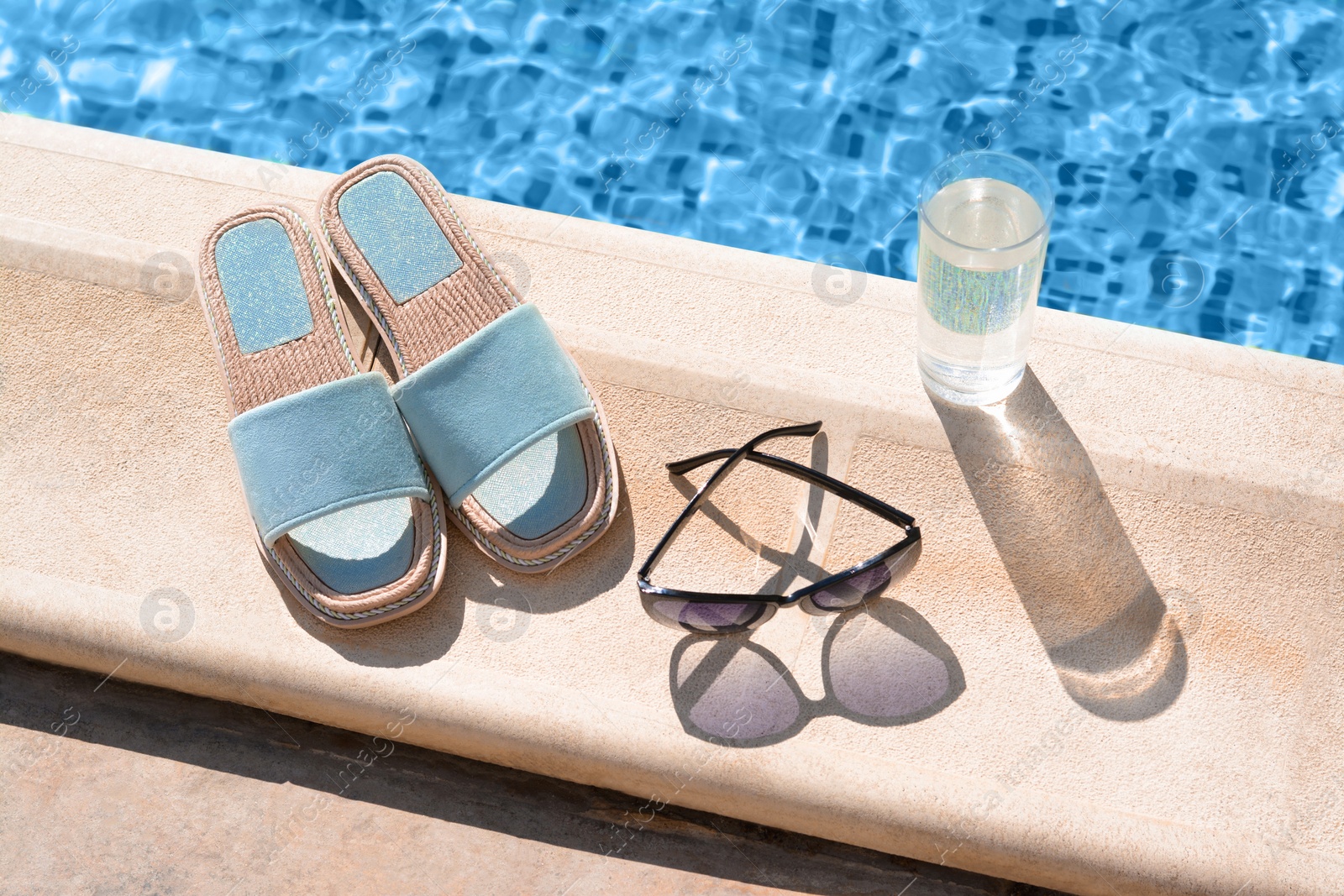 Photo of Stylish sunglasses, slippers and glass of water at poolside on sunny day. Beach accessories