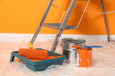 Photo of Ladder, cans of paint and renovation equipment on floor near orange wall indoors