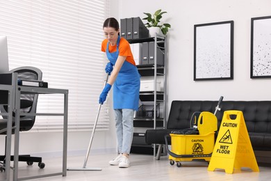 Photo of Cleaning service worker washing floor with mop, low angle view. Bucket with wet floor sign in office