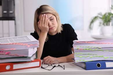 Photo of Overwhelmed woman sitting at table with stacks of documents and folders in office