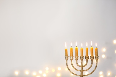 Photo of Golden menorah with burning candles against light grey background and blurred festive lights, space for text