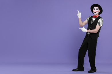 Photo of Mime artist in hat pointing at something on purple background. Space for text