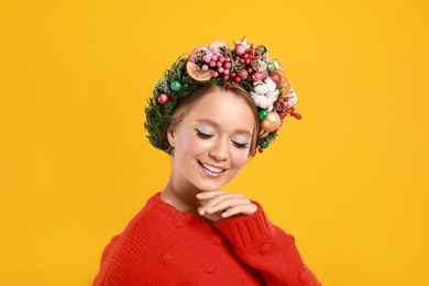 Beautiful young woman wearing Christmas wreath on yellow background