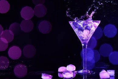 Photo of Glass of martini with ice and splashes in neon lights on dark background, bokeh effect. Space for text