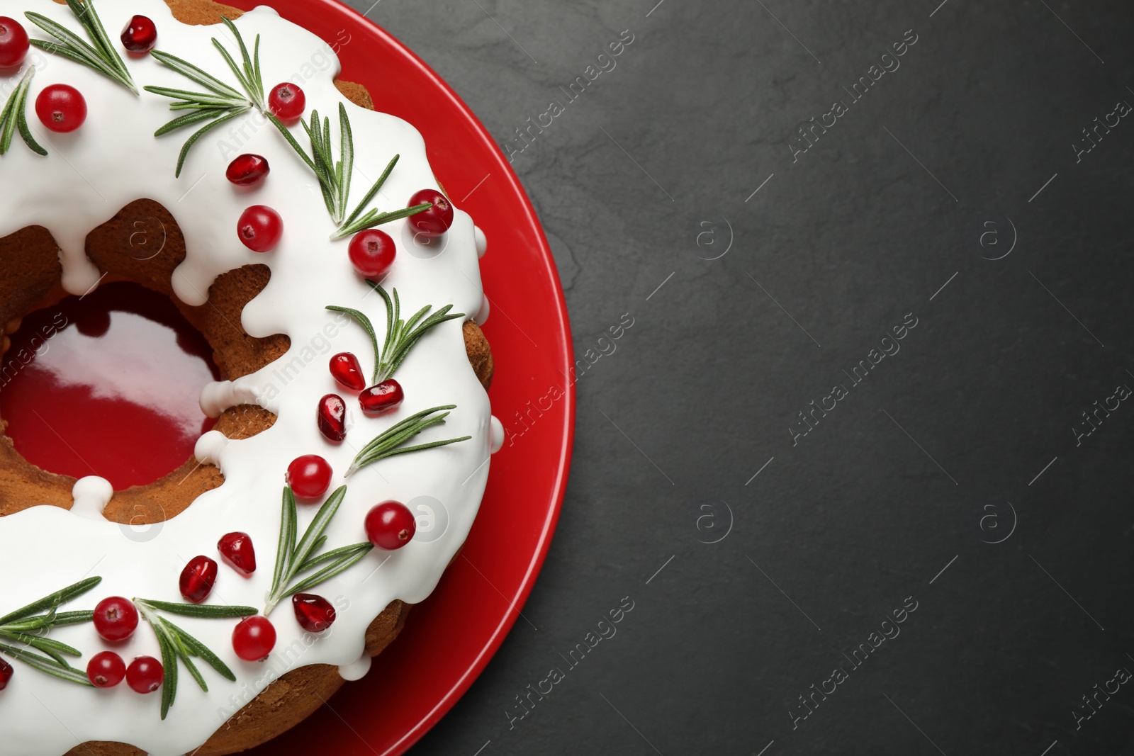 Photo of Traditional Christmas cake decorated with glaze, pomegranate seeds, cranberries and rosemary on dark grey table, top view. Space for text