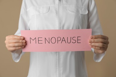 Doctor holding paper sheet with word MENOPAUSE on beige background, closeup