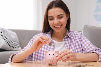 Photo of Happy young woman putting coin into piggy bank at home