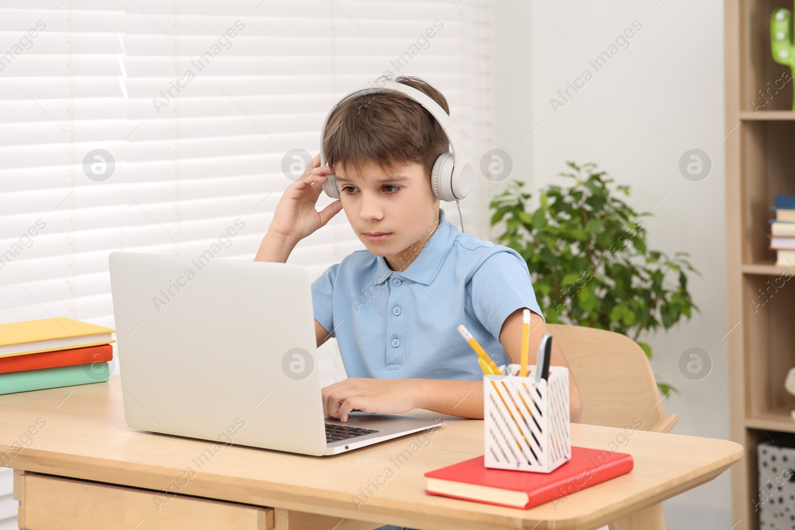 Photo of Boy using laptop and headphones at desk in room. Home workplace