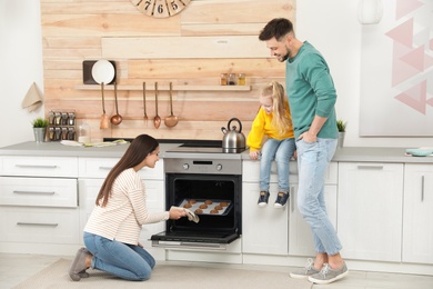 Photo of Woman and her family taking out tray with baked cookies from oven in kitchen