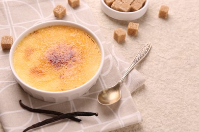 Photo of Delicious creme brulee in bowl, vanilla pods, sugar cubes and spoon on light textured table, closeup