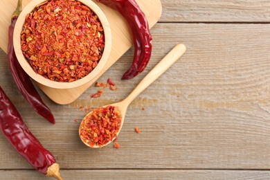 Photo of Aromatic spice. Red chili pepper flakes in bowl, spoon and pods on wooden table, flat lay. Space for text