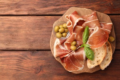 Photo of Slices of tasty cured ham, olives, bread and basil on wooden table, top view. Space for text
