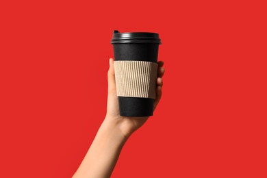 Photo of Woman holding takeaway paper coffee cup with cardboard sleeve on red background, closeup