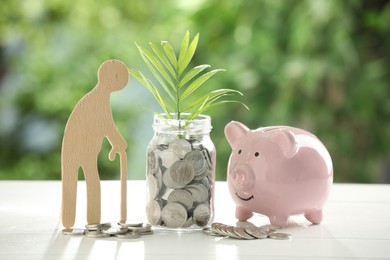 Pension savings. Figure of senior man, piggy bank, jar with coins and twig on white table against blurred green background