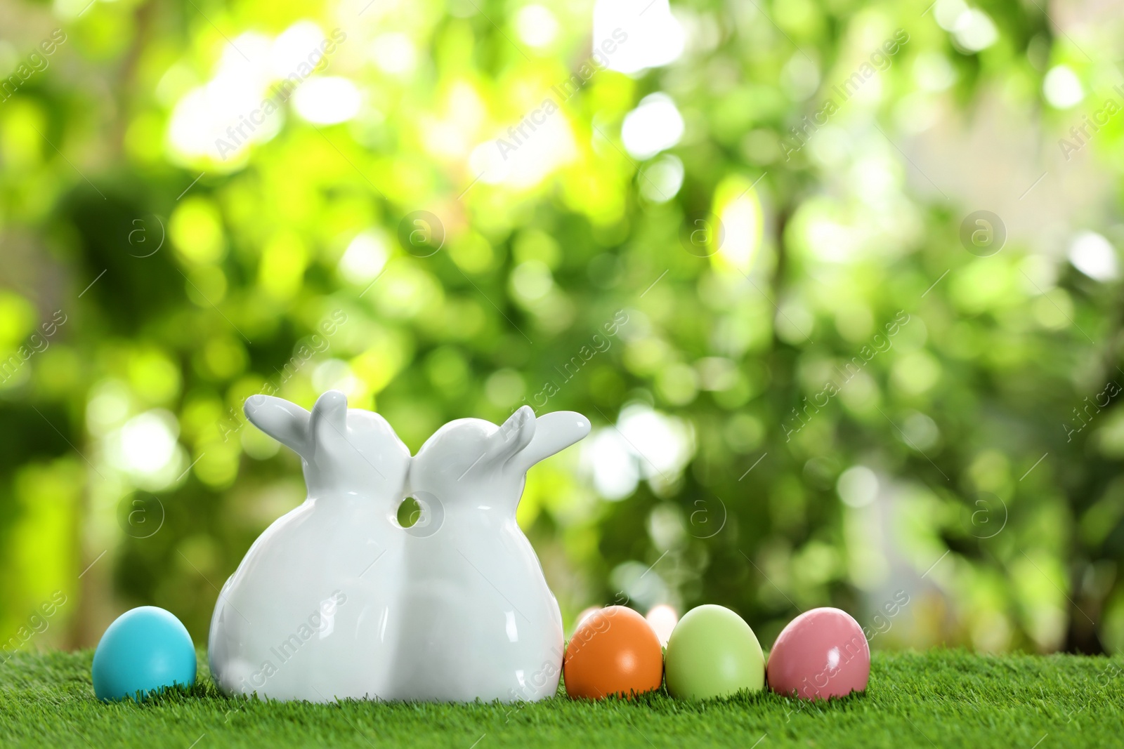 Photo of Cute ceramic Easter bunnies and dyed eggs on green grass against blurred background, space for text