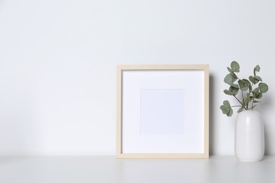 Empty photo frame and vase with decorative eucalyptus leaves on white table. Mockup for design