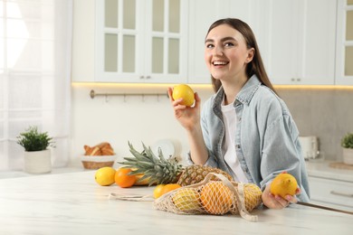 Photo of Woman with lemons and string bag of fresh fruits at light marble table in kitchen