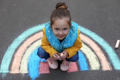 Happy child near chalk drawing of rainbow on asphalt, above view