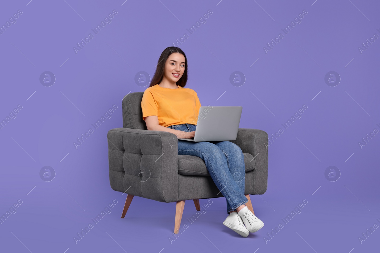 Photo of Smiling young woman with laptop sitting in armchair on lilac background, space for text