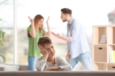 Little unhappy boy sitting on sofa while parents arguing at home