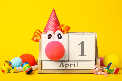 Photo of Wooden block calendar with clown face and party decor on yellow background. April fool's day