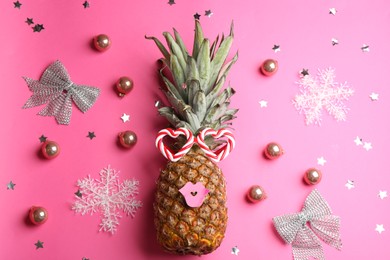 Pineapple with funny glasses and Christmas decor on pink background, flat lay. Creative concept